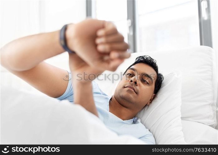 people, bedtime and rest concept - indian man looking at health tracker in bed at home. indian man with health tracker in bed at home
