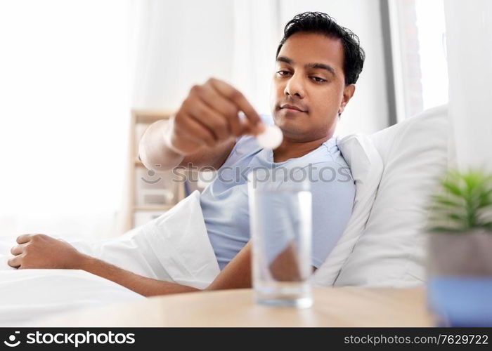 people, bedtime and rest concept - indian man lying in bed and dropping medicine pill into glass of water at home. man in bed dropping medicine into glass of water