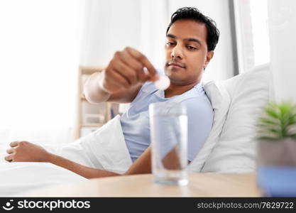 people, bedtime and rest concept - indian man lying in bed and dropping medicine pill into glass of water at home. man in bed dropping medicine into glass of water