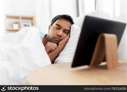 people, bedtime and rest concept - indian man sleeping in bed and tablet pc computer on table at home. indian man sleeping in bed and tablet pc at home