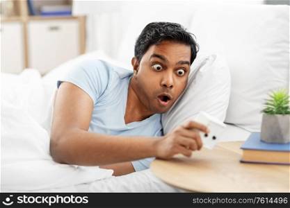 people, bedtime and rest concept - overslept indian man looking at alarm clock lying in bed at home. overslept indian man in bed looking at alarm clock