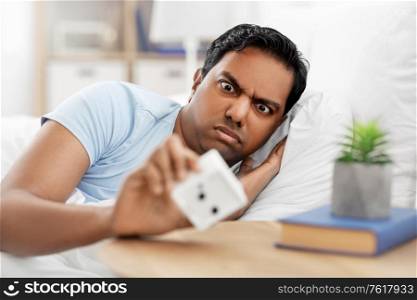 people, bedtime and rest concept - overslept indian man looking at alarm clock lying in bed at home. overslept indian man in bed looking at alarm clock