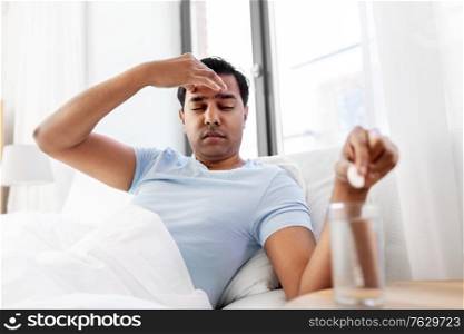 people, bedtime and rest concept - sick indian man lying in bed and dropping medicine pill into glass of water at home. sick man in bed with medicine and glass of water