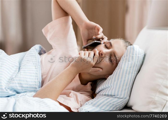 people, bedtime and rest concept - sleepy girl calling on smartphone lying in bed at home. sleepy girl calling on smartphone in bed at home