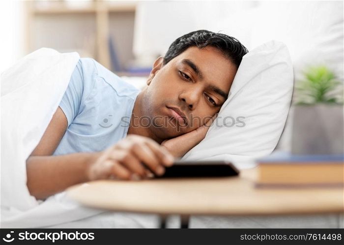 people, bedtime and rest concept - sleepy indian man in bed looking at smartphone lying on table at home. sleepy indian man in bed looking at smartphone