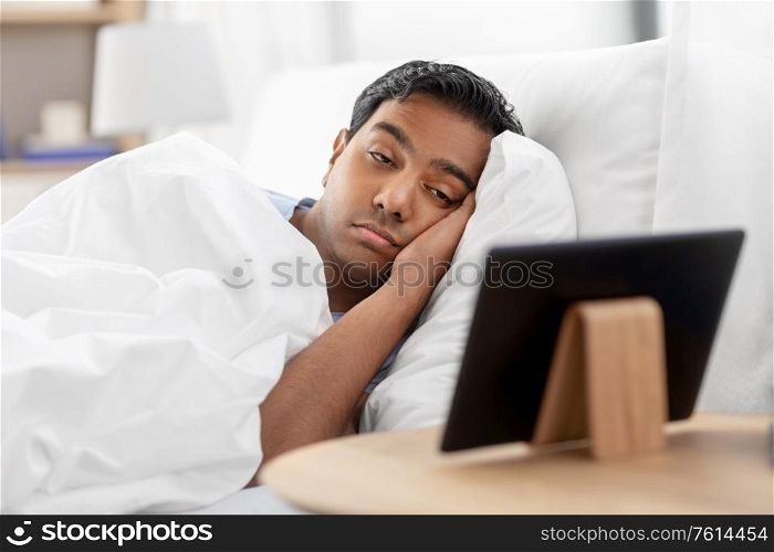 people, bedtime and rest concept - sleepy indian man looking at tablet pc computer lying in bed at home. sleepy indian man looking at tablet pc in bed