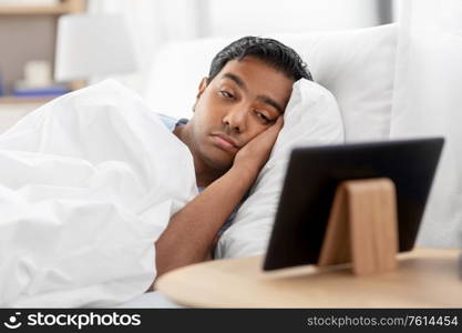 people, bedtime and rest concept - sleepy indian man looking at tablet pc computer lying in bed at home. sleepy indian man looking at tablet pc in bed