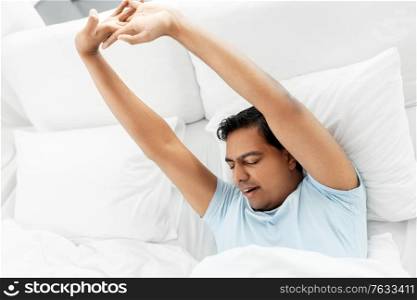 people, bedtime and rest concept - sleepy indian man stretching in bed at home. sleepy indian man stretching in bed at home