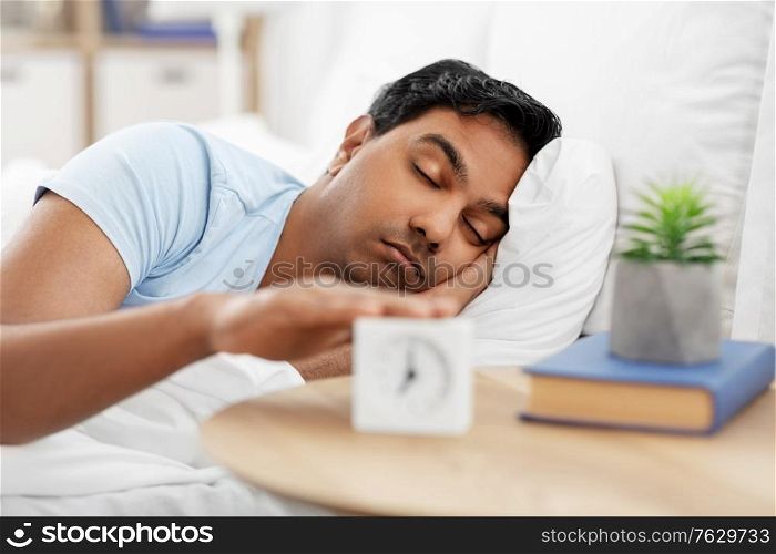 people, bedtime and rest concept - sleepy indian man with alarm clock lying in bed at home. sleepy indian man with alarm clock lying in bed