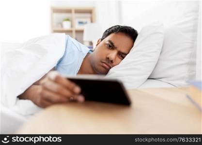 people, bedtime and rest concept - sleepy indian man with smartphone lying in bed at home. sleepy indian man with smartphone lying in bed