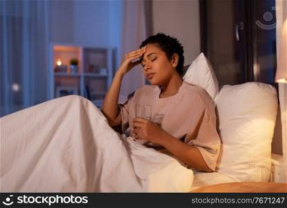 people, bedtime and rest concept - stressed african american woman having headache and drinking water in bed at night. stressed african woman lying in bed at night