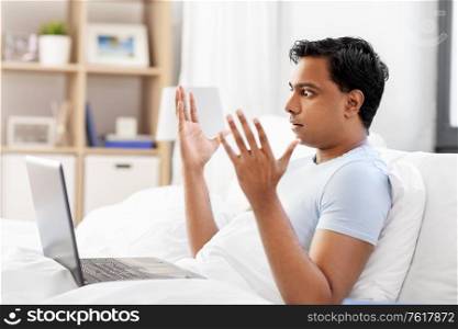 people, bedtime and rest concept - stressed indian man with laptop computer lying in bed at home. stressed indian man with laptop in bed at home
