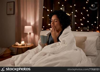 people, bedtime and rest concept - tired sleepy woman in pajamas wrapped in blanket with coffee yawning in bed at night. tired woman with coffee yawning in bed at night