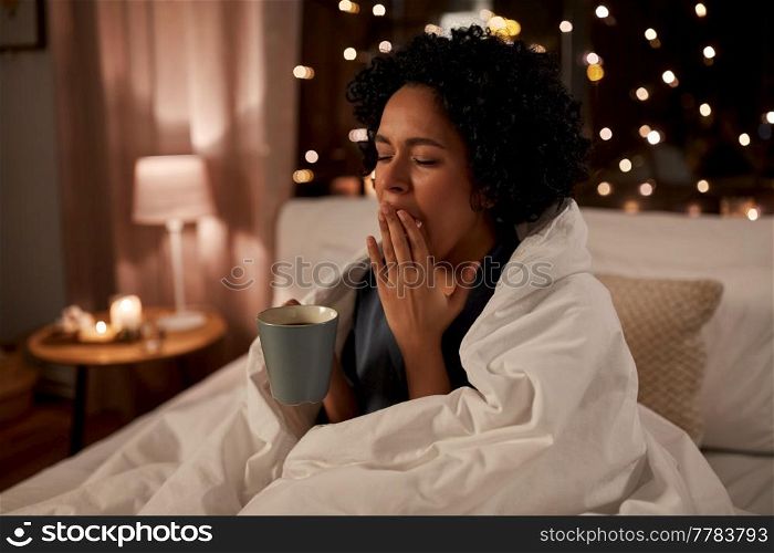 people, bedtime and rest concept - tired sleepy woman in pajamas wrapped in blanket with coffee yawning in bed at night. tired woman with coffee yawning in bed at night