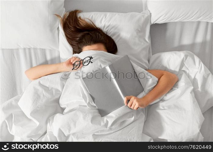 people, bedtime and rest concept - woman lying in bed under white blanket or duvet with book and glasses. woman lying in bed with book and glasses