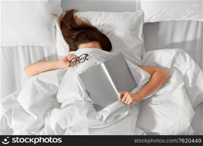 people, bedtime and rest concept - woman lying in bed under white blanket or duvet with book and glasses. woman lying in bed with book and glasses
