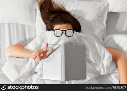 people, bedtime and rest concept - woman lying in bed under white blanket or duvet with book and glasses showing peace hand sign. woman lying in bed with book and glasses