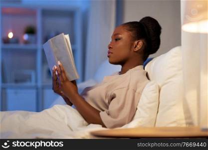 people, bedtime and rest concept - young african american woman reading book in bed at home at night. young woman reading book in bed at home