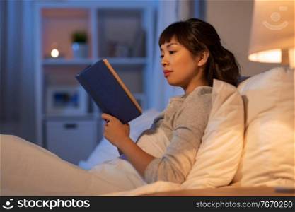 people, bedtime and rest concept - young asian woman reading book in bed at home at night. young asian woman reading book in bed at home