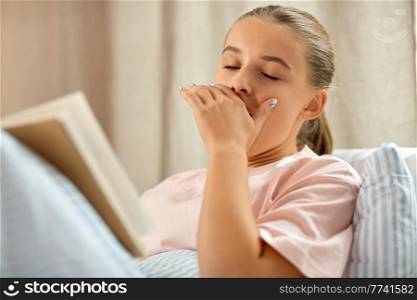 people, bedtime and sleeping concept - yawning girl reading book in bed at home. yawning girl reading book in bed at home