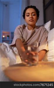 people, bedtime and thirst concept - african american woman drinking water in bed at night. african american woman drinking water at night