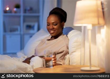 people, bedtime and thirst concept - happy smiling african american woman drinking water at home at night. african woman drinking water in bed at night