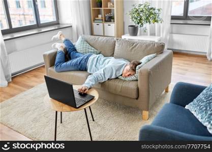 people, boredom and depression concept - bored or lazy young man with laptop computer lying on sofa at home. bored man with laptop lying on sofa at home