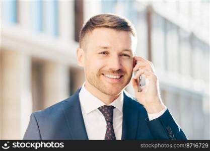 People, business and career concept. Handsome cheerful successful male in formal clothes, has bristle and positive smile, speaks via smart phone with businessman, poses against blurred background