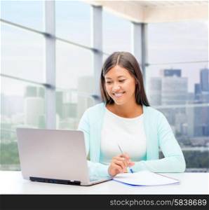 people, business and technology concept - smiling woman in with laptop computer and notebook sitting at table over office window background