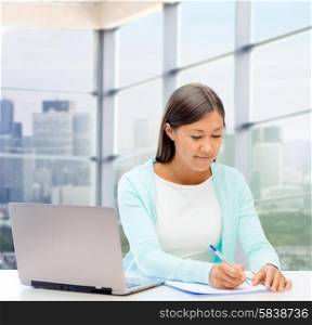people, business and technology concept - smiling woman in with laptop computer writing to notebook over office window background