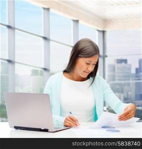 people, business and technology concept - smiling woman with laptop computer sitting at table and reading papers over office window background