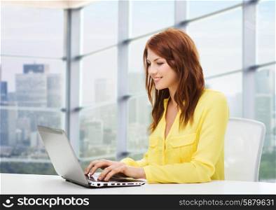 people, business and technology concept - smiling young woman with laptop computer sitting at table over office window background