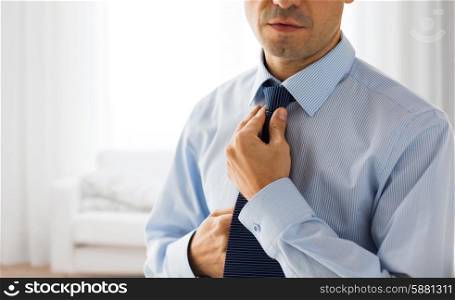 people, business, fashion and clothing concept - close up of man in shirt dressing up and adjusting tie on neck over living room background