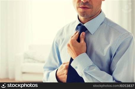 people, business, fashion and clothing concept - close up of man in shirt dressing up and adjusting tie on neck over living room background