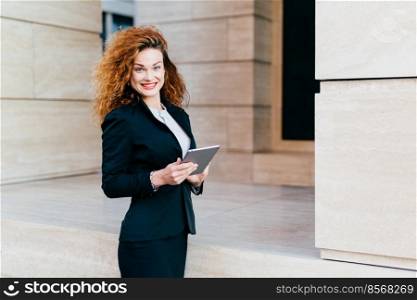 People, business, innovations and technology concept. Happy curly beautiful woman with red painted lips, wearing formal clothes, holding modern tablet computer, posing against her office builduing