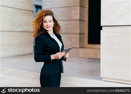 People, business, modern technology concept. Pretty businesswoman wearing black jacket and skirt standing near her office with modern tablet, developing business strategy, looking into camera