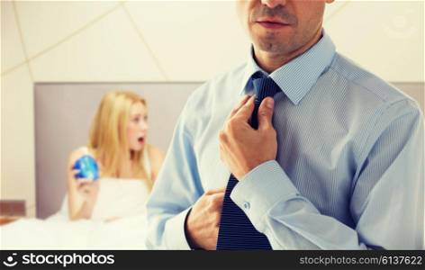 people, business, morning and work concept - close up of man in shirt dressing up and adjusting tie on neck over bedroom with woman holding alarm clock background