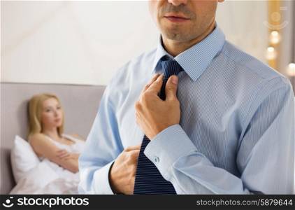 people, business, relations and problems concept - close up of man in shirt dressing up and adjusting tie on neck over woman in bed background