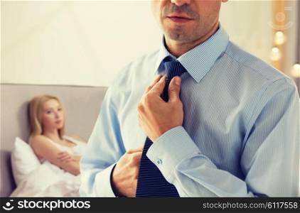 people, business, relations and problems concept - close up of man in shirt dressing up and adjusting tie on neck over woman in bed background