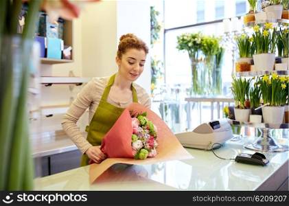 people, business, sale and floristry concept - happy smiling florist woman wrapping bunch into paper at flower shop