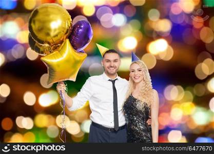 people, celebration and holidays concept - happy couple with party caps and balloons over festive lights background. happy couple with balloons over party lights. happy couple with balloons over party lights