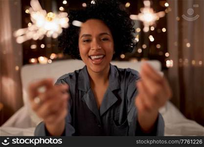 people, celebration and holidays concept - happy smiling woman in pajamas with sparklers sitting in bed at night. happy woman with sparklers sitting in bed at night