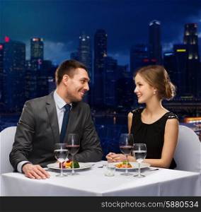 people, celebration, romantic and holidays concept - smiling couple with red wine and food talking at restaurant over night city background