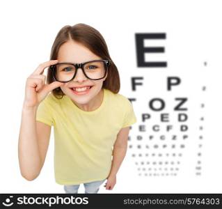 people, childhood, healthcare and vision concept - smiling little girl in black eyeglasses over white background with eye chart