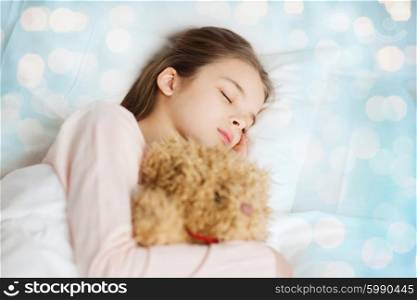 people, childhood, rest and comfort concept - girl sleeping with teddy bear toy in bed over blue lights background