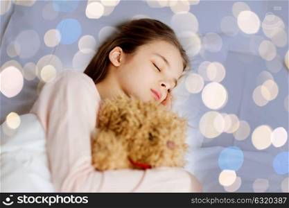 people, childhood, rest and comfort concept - girl sleeping with teddy bear toy in bed over holidays lights background. girl sleeping with teddy bear toy in bed