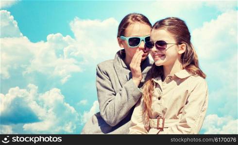 people, children and friendship concept - happy little girl in sunglasses whispering her secret to friends ear or gossiping over blue sky and clouds background