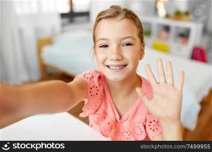 people, children and technology concept - girl taking selfie and waving hand at home. happy girl taking selfie and waving hand at home