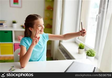people, children and technology concept - girl with laptop computer and smartphone taking selfie at home and showing peace hand sign. happy girl with smartphone taking selfie at home