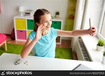 people, children and technology concept - girl with laptop computer and smartphone taking selfie at home and waving hand. happy girl with smartphone taking selfie at home